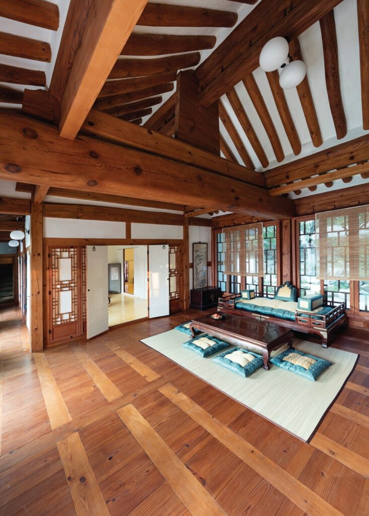 서울 북촌 백인제 가옥의 안방 대청마루 전경. 천장을 가로지르는 웅장한 대들보와 서까래는 당대 최고급 자재인 압록강 흑송의 위용을 보여준다. 넓게 펼쳐진 우물마루 위에는 전통 보료와 탁자가 놓여 있으며, 측면의 복도와 유리 창호는 전통 공간에 근대적 생활 양식이 접목되었음을 나타낸다.