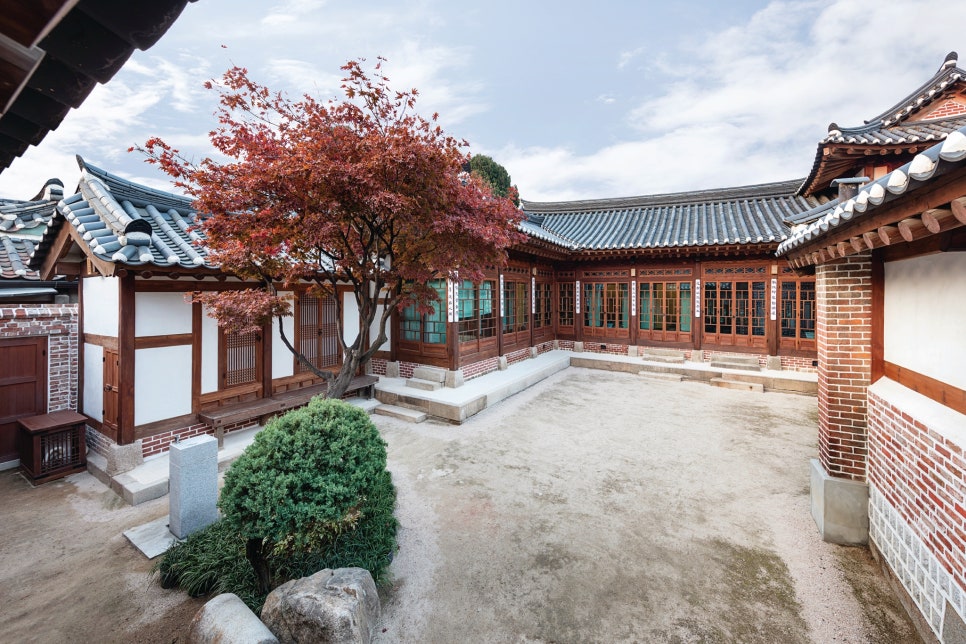 서울 북촌 백인제 가옥의 안채 마당 전경. 'ㄱ'자형 한옥 건물에 둘러싸인 정갈한 마당 한가운데 붉게 물든 단풍나무가 서 있다. 전통적인 기와지붕 아래로 근대식 유리 미서기문과 붉은 벽돌이 사용된 벽면이 보여, 개화기 상류층 가옥의 독특한 건축 양식을 확인할 수 있다.