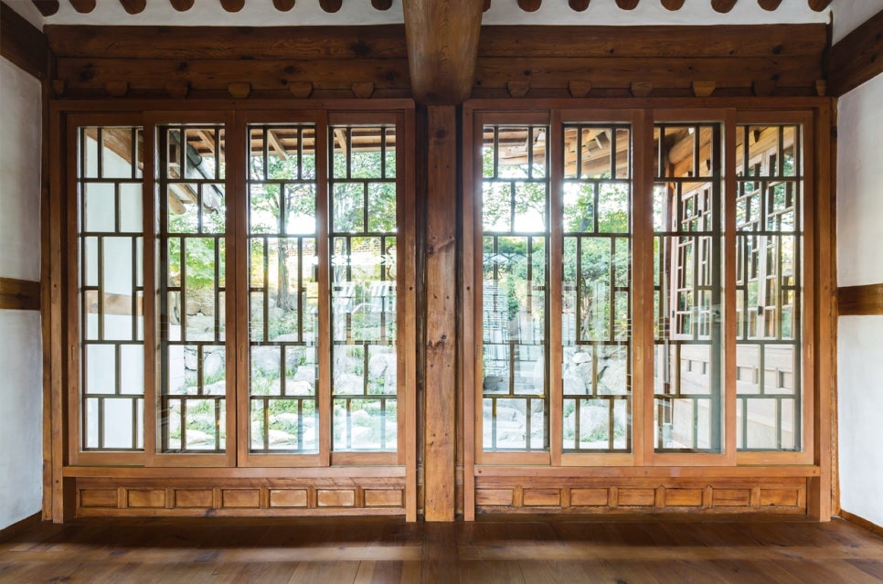 서울 북촌 백인제 가옥 안채 내부에서 유리창 너머로 바라본 뒷마당 풍경. 전통적인 우물마루와 나무 기둥 사이에 설치된 미닫이문은 창호지 대신 투명한 유리를 사용하여 개방감을 주었다. 유리 너머로 보이는 돌담과 초록의 정원 풍경이 실내로 그대로 들어오며, 단열과 조망을 동시에 고려한 근대 한옥의 실용적인 미학을 보여준다.