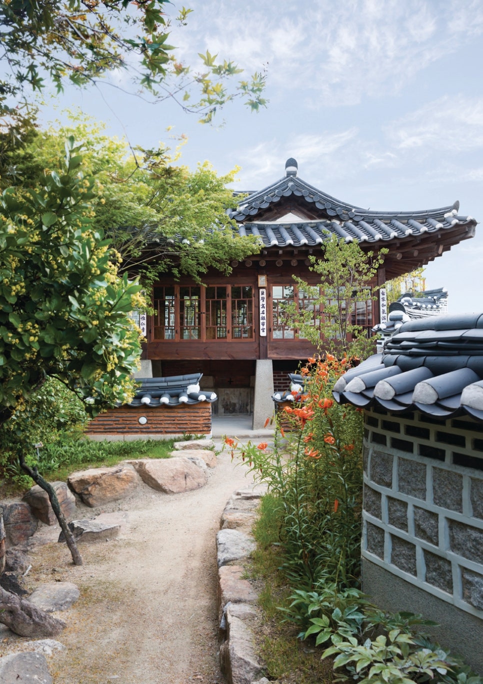 서울 북촌 백인제 가옥의 가장 높은 곳에 위치한 별당채의 전경. 전통 한옥에서는 보기 드문 2층 누각 형식의 건물로, 전면의 유리창과 나무 널문이 근대적 특징을 보여준다. 건물 주변을 감싼 푸른 단풍나무와 붉은 꽃들이 어우러져, 북촌 일대를 조망할 수 있는 휴식처로서의 고즈넉하고 은밀한 분위기를 자아낸다.