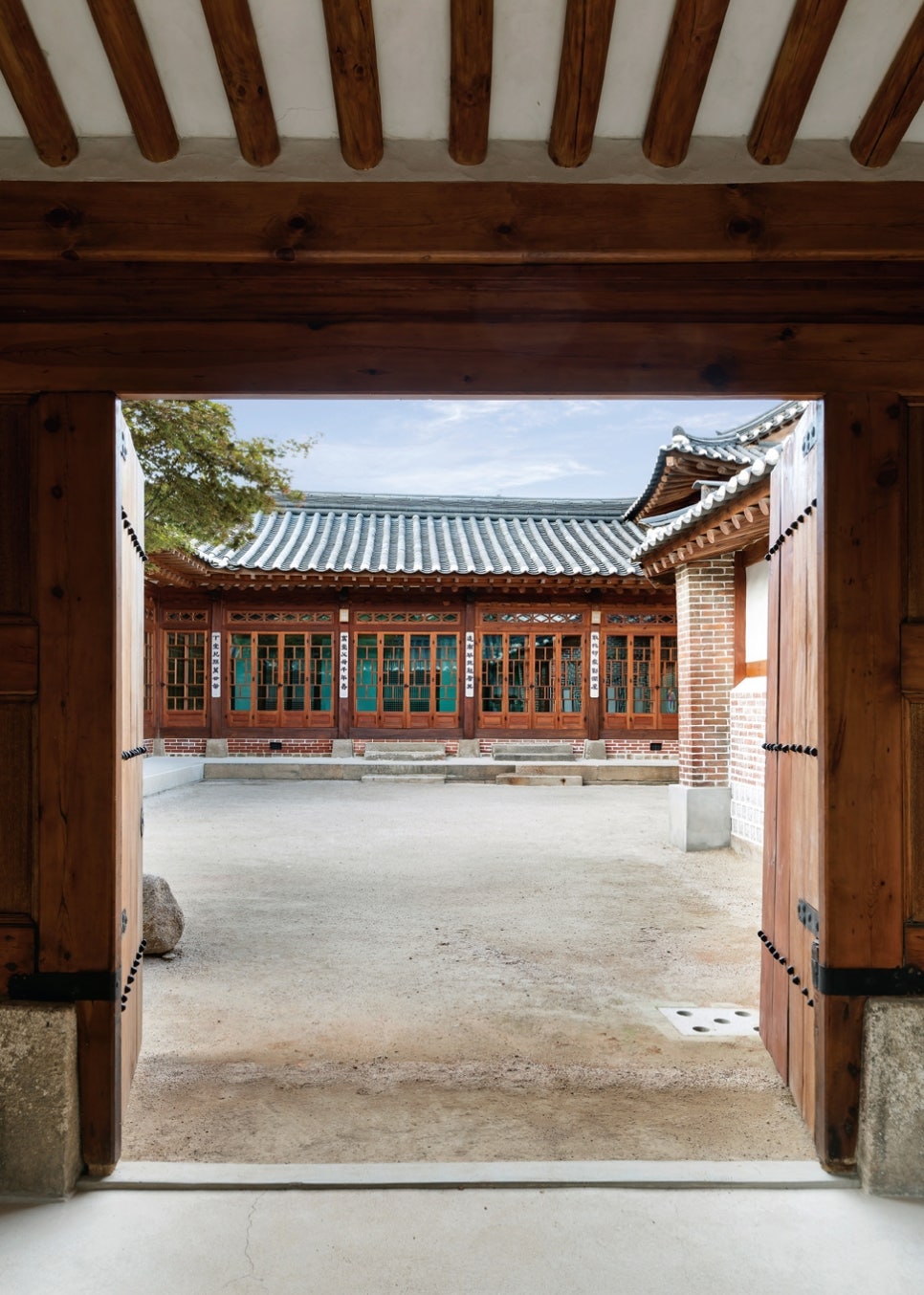 서울 북촌 백인제 가옥의 중문채(내부 대문)가 활짝 열려 있는 모습. 두꺼운 나무 대문이 만들어낸 프레임 너머로 정갈한 안채 마당과 유리 미서기문이 설치된 안채 건물이 보인다. 우측의 붉은 벽돌 벽과 전통 목조 대문이 어우러져, 사랑채(외부인 공간)와 안채(가족 공간)를 구분 짓는 공간적 경계를 시각적으로 보여준다.