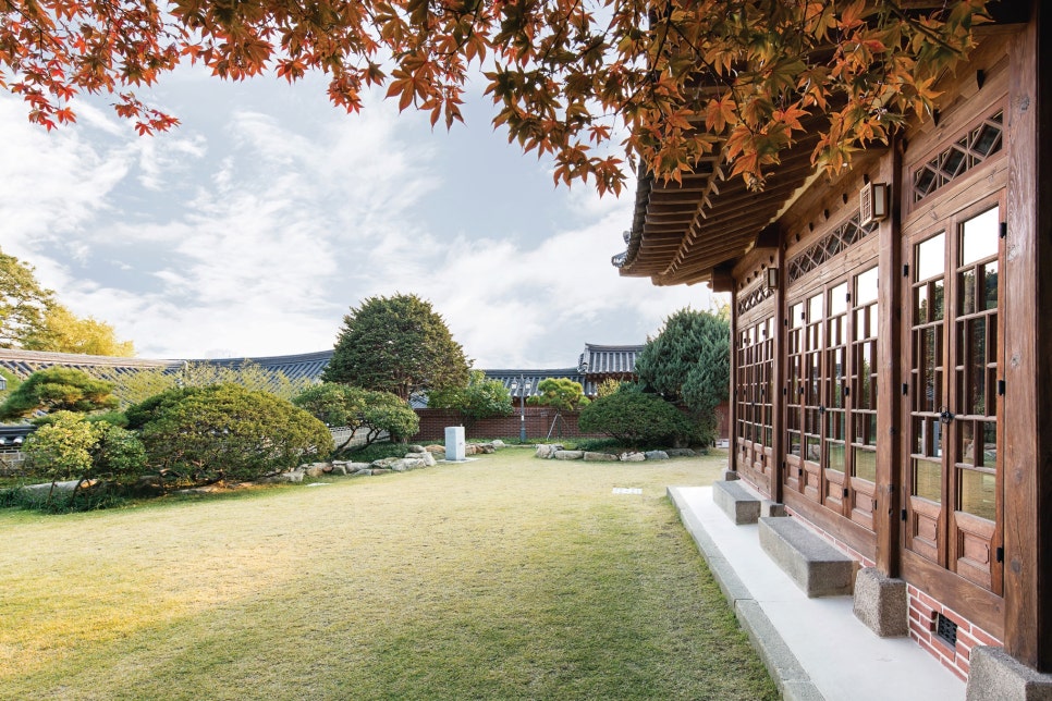 서울 북촌 백인제 가옥의 사랑채와 넓은 정원 전경. 붉은 단풍나무 가지 아래로 잘 가꿔진 푸른 잔디밭과 조경석이 보이며, 우측에는 전면이 유리창으로 된 독특한 양식의 사랑채 건물이 자리하고 있다. 일제강점기 상류층의 사교 모임과 연회를 위해 설계된 근대 한옥의 개방적인 구조를 잘 보여준다.