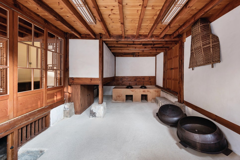 서울 북촌 백인제 가옥의 부엌 내부 전경. 흰 회벽과 나무 서까래 아래로 전통 방식의 아궁이와 무쇠 가마솥이 놓여 있다. 특이하게도 부엌과 방을 연결하는 문이 전통 창호가 아닌 근대식 유리 미서기문으로 되어 있어, 개항기 이후 변화된 한옥의 생활 양식을 보여준다.