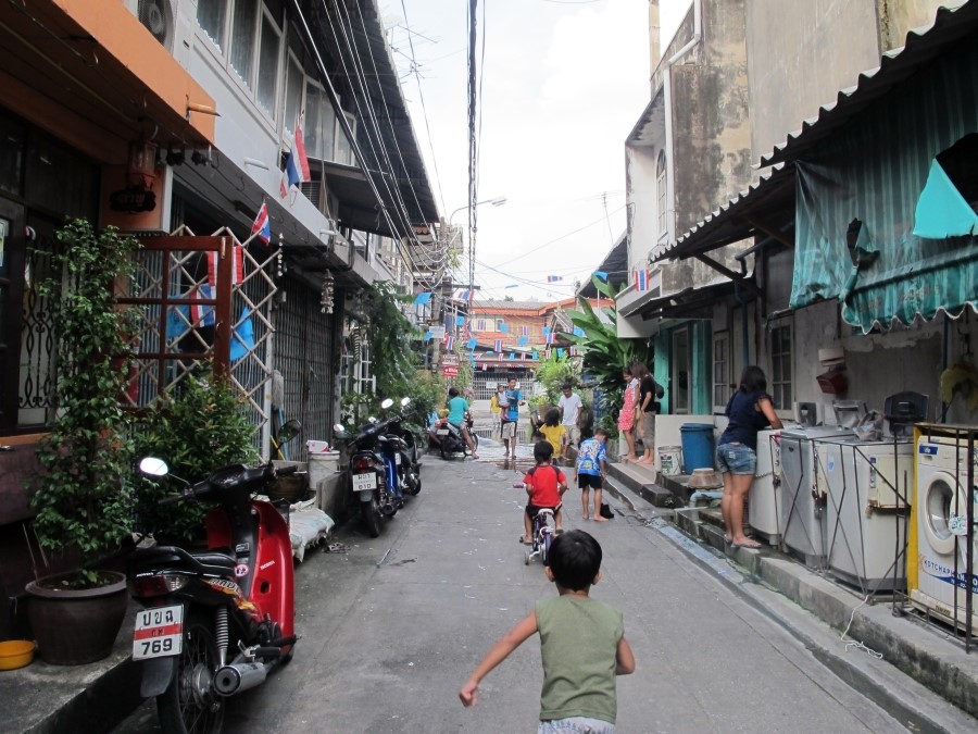 방콕의 한적한 주택가 골목길. 아이들이 자전거를 타거나 뛰어놀고 있고, 노상에 세탁기와 오토바이가 줄지어 있는 풍경
