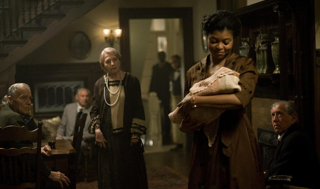 영화 벤자민 버튼의 한 장면. 양로원 복도에서 퀴니가 노인의 얼굴을 한 아기 벤자민을 담요에 싸서 소중하게 안고 있다. 배경에 있는 다른 노인들과 신사는 기이한 모습의 아기를 낯선 눈빛으로 쳐다보고 있다.