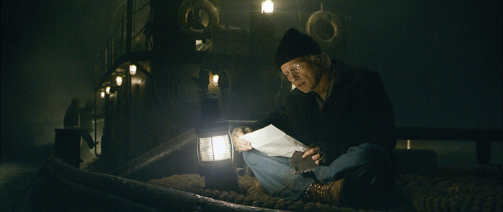 영화 의 한 장면. 어두운 밤, 예인선 갑판에 홀로 앉아 램프의 희미한 불빛에 의지해 편지를 읽고 있는 노년 외모의 벤자민 버튼. 밧줄이 놓인 거친 작업 환경과 벤자민의 고요하고 진지한 표정이 대조를 이루며, 고독한 삶 속에서도 타인과의 연결을 놓지 않으려는 내면을 보여준다.