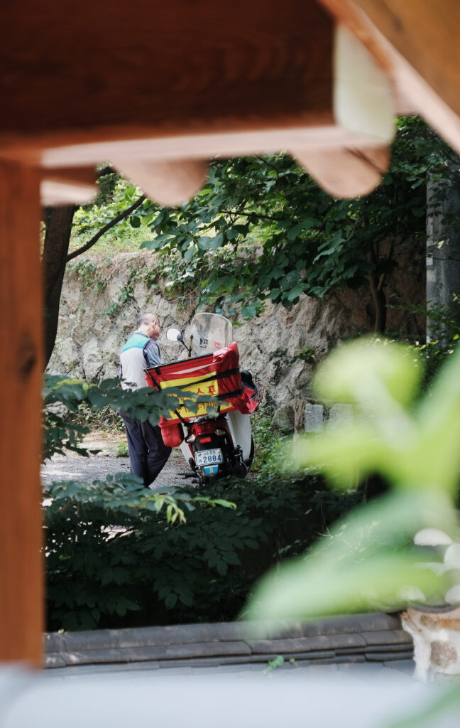 돌담과 초록빛 수풀이 우거진 좁은 골목길에서 오토바이를 세우고 우편물을 확인하는 우편배달부의 뒷모습