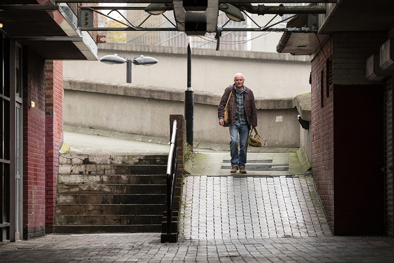 영화 의 한 장면. 회색빛 콘크리트 건물과 차가운 철골 구조물 사이로 난 경사로를 홀로 걸어 올라가는 다니엘의 모습. 낡은 가방을 메고 다소 지친 기색이지만 묵묵히 발걸음을 옮기는 장면은, 거대한 시스템 안에서 고립된 개인이 겪는 외로운 싸움과 무게감을 상징함.