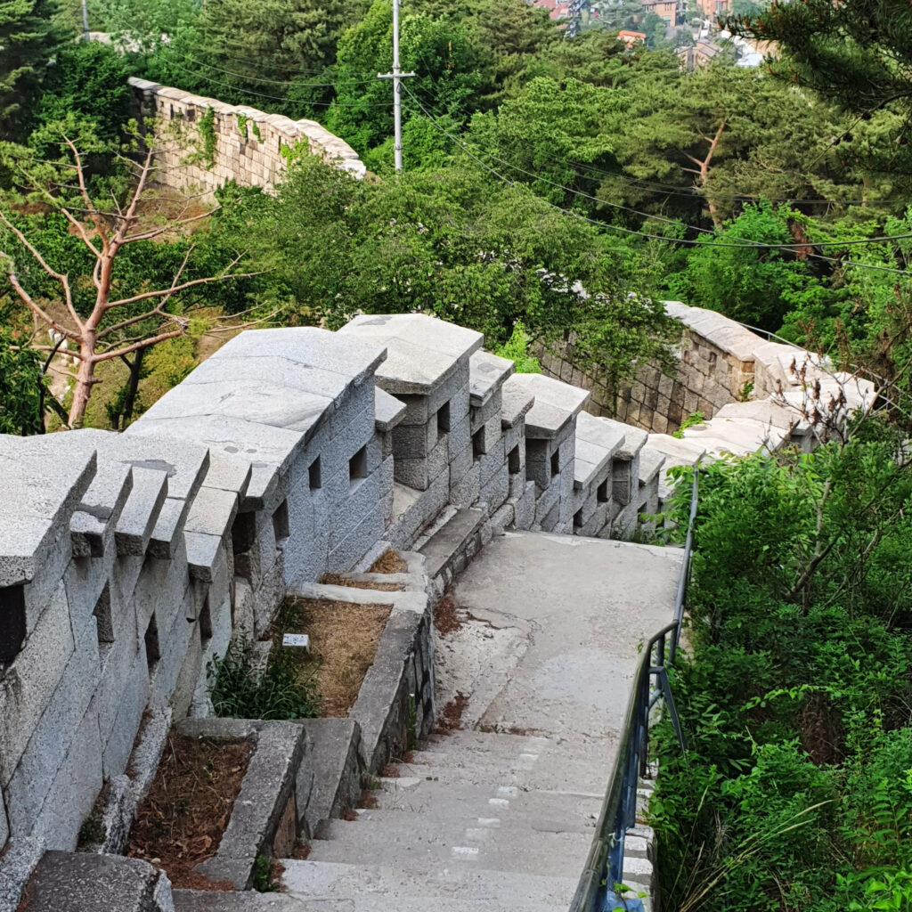 산등성이를 따라 굽이굽이 이어지는 견고한 조선시대 성곽 돌담과 그 주변을 감싸는 푸른 나무들, 멀리 내려다보이는 서울 도심의 전경