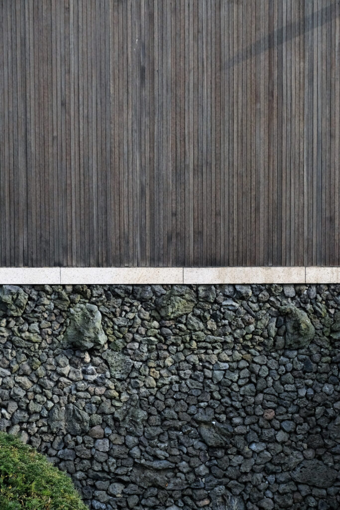 제주 방주교회의 외벽 건축 자재 디테일. 상단은 세로결이 살아있는 짙은 색 목재로 마감되었고, 하단은 제주 고유의 거친 현무암을 쌓아 올린 돌담으로 구성되어 있다. 서로 다른 물성을 지닌 나무와 돌이 만나 조화를 이루며, 자연과 동화되려는 이타미 준의 건축 철학과 제주의 토착성을 보여준다.