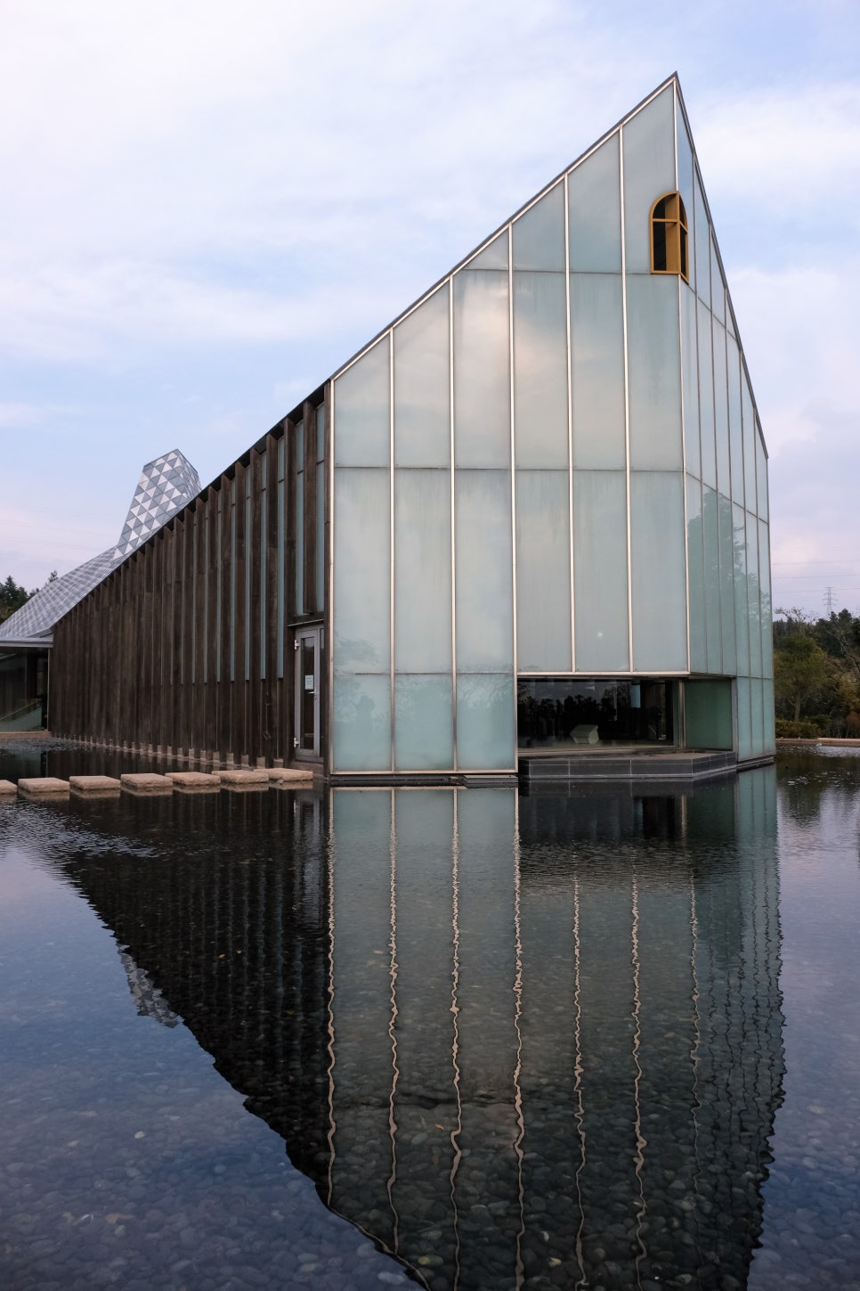 제주 방주교회의 정면 유리 벽면(Facade)과 수면 위 반영. 하늘 높이 솟은 투명한 유리창과 수직 프레임이 잔잔한 물에 거울처럼 비치며, 마치 물살을 가르는 방주의 앞머리(선수) 형상을 하고 있다. 물 밑의 자갈과 유리의 투명함이 어우러져 이타미 준 건축 특유의 투명성과 조형미를 보여준다.