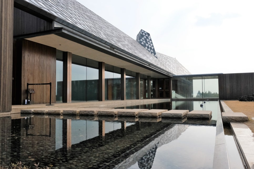 제주 방주교회 건물 측면을 감싸고 있는 잔잔한 수조(Reflection Pool)와 그 위를 건너는 네모난 돌 징검다리 전경. 투명한 유리 벽과 징크 소재의 기하학적 지붕 패턴이 물 위에 거울처럼 비치며, 마치 건물이 물 위에 떠 있는 듯한 '노아의 방주' 형상을 극적으로 보여준다.