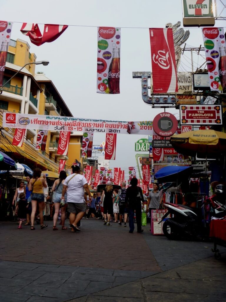 낮 시간대 방콕 카오산 로드의 번잡한 거리. 코카콜라 깃발과 다양한 언어의 현수막이 걸려 있고 여행자들이 거리를 걷고 있는 풍경