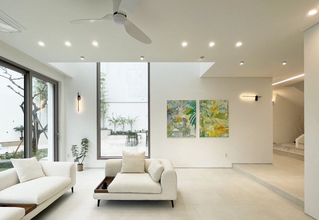 묵화담 1층 거실 인테리어 전경: 화이트 패브릭 소파와 실링팬, 그리고 전은숙 작가의 연작 회화가 대나무 중정 뷰와 어우러진 갤러리형 공간