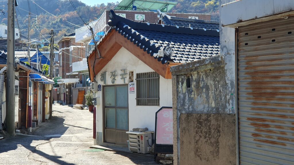 목포 원도심의 한적한 골목길 풍경. 코너에 위치한 작은 식당은 검은색 기와지붕 아래 '해장국'이라는 간판을 달고 있어 소박한 정취를 풍긴다. 주변의 녹슨 셔터와 낡은 담벼락, 좁은 도로가 어우러져 시간이 멈춘 듯한 목포 특유의 레트로한 골목 감성을 보여준다.