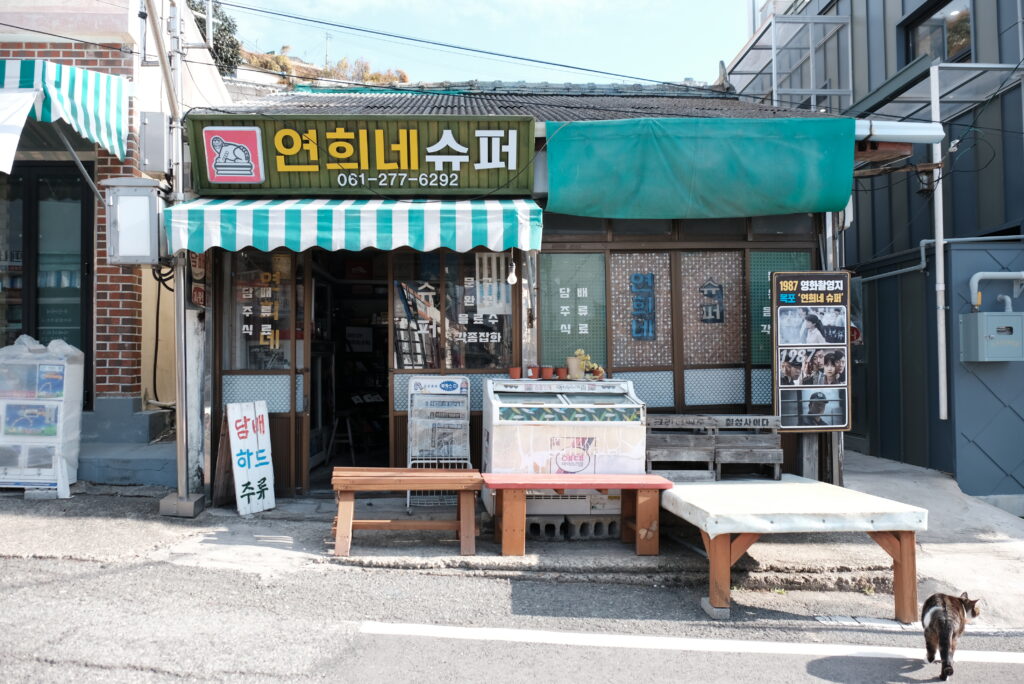 목포 서산동 시화골목에 위치한 '연희네슈퍼' 전경. 영화 촬영지로 유명한 이곳은 초록색 줄무늬 천막, 낡은 미닫이문, 가게 앞 평상 등 1980년대 구멍가게의 모습을 완벽하게 재현하고 있다. 우측에는 영화 포스터가 세워져 있어 레트로한 감성을 더한다.