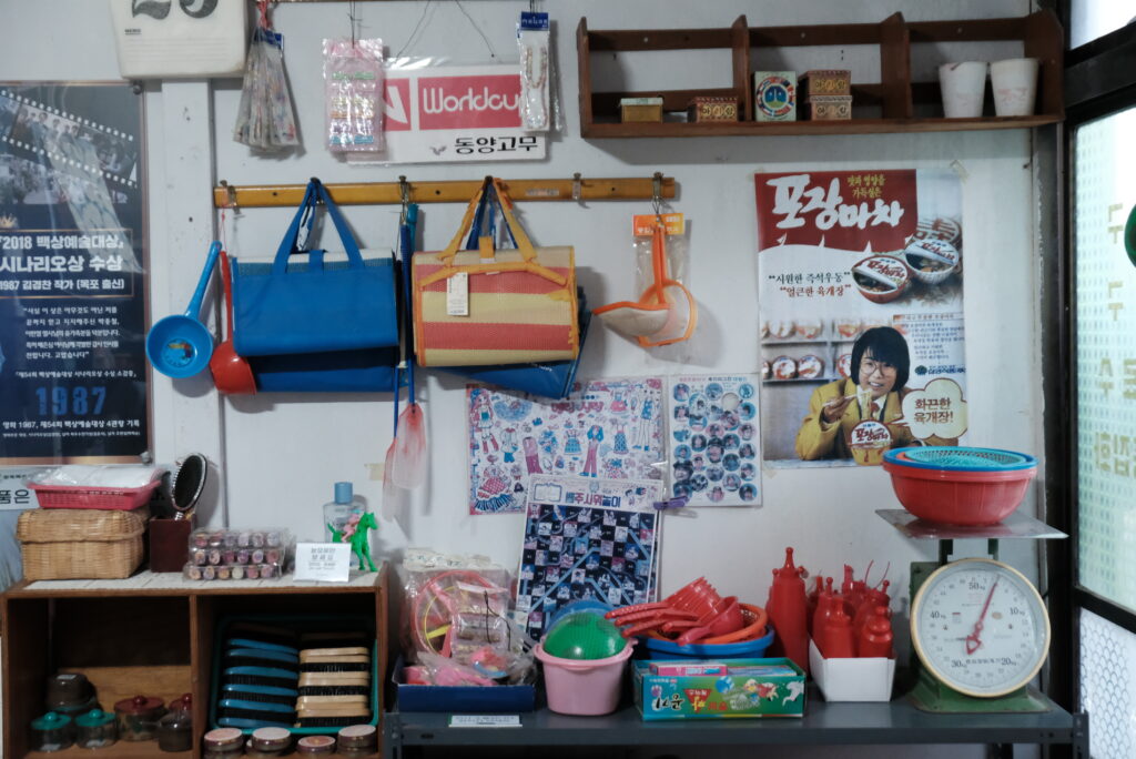 목포 연희네슈퍼 내부 벽면에 진열된 1980년대 생활용품들. 빛바랜 '포장마차' 우동 광고 포스터와 '월드컵' 고무신 간판 아래로, 알록달록한 나일론 장바구니와 플라스틱 바가지, 옛날 저울이 놓여 있다. 좌측에는 영화 의 백상예술대상 수상 기념 포스터가 부착되어 있어 촬영지로서의 의미를 더한다.