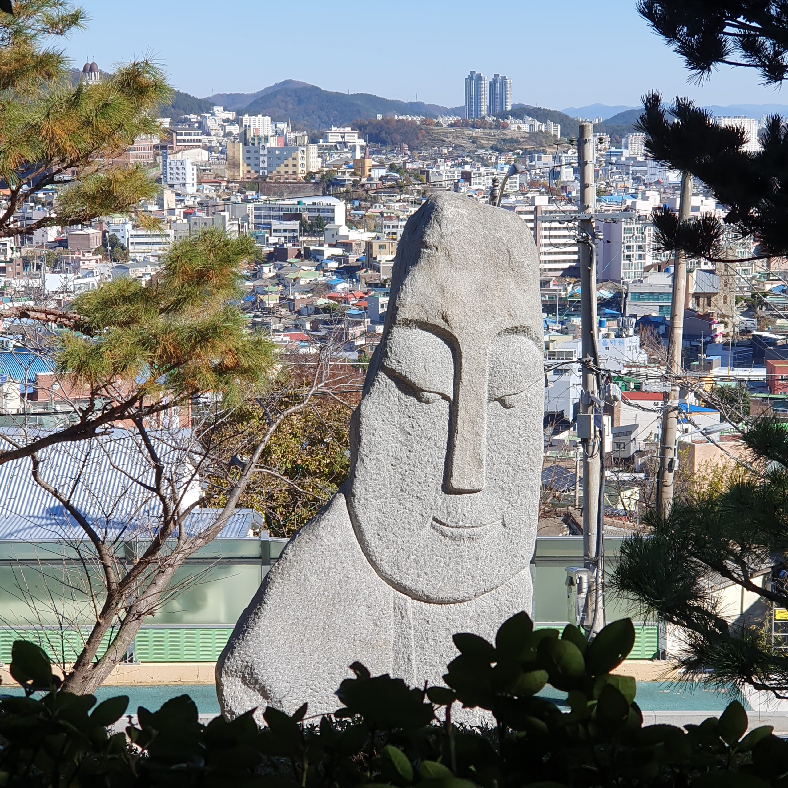 목포 유달산 조각공원에 설치된 거대한 석조 조각상. 온화한 표정으로 눈을 감고 있는 사람 얼굴 형상의 조각상 뒤로, 목포 원도심의 빽빽한 건물들과 스카이라인이 한눈에 들어온다. 예술 작품과 도시 풍경, 그리고 소나무가 어우러져 목포 특유의 평화로운 분위기를 자아낸다.