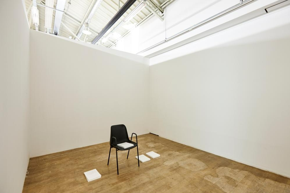 하얀 벽의 미니멀한 공간 한가운데 검은 의자가 놓여 있고 주변에 흰 책들이 배치된 참여형 전시 공간.