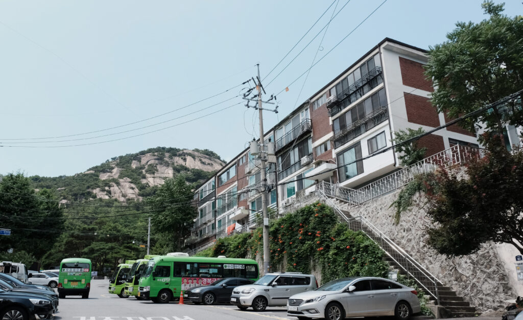 인왕산의 웅장한 암벽 아래 초록색 09번 마을버스가 주차되어 있고, 그 위로 붉은 벽돌의 옥인빌라가 자리한 수성동계곡 입구 전경