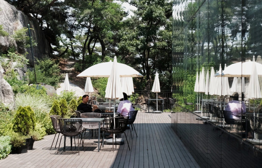 인왕산의 거대한 바위와 숲을 배경으로, 하얀 파라솔 아래 테이블에 앉아 휴식을 취하는 사람들과 숲의 녹음을 반사하는 통유리 건물의 외관