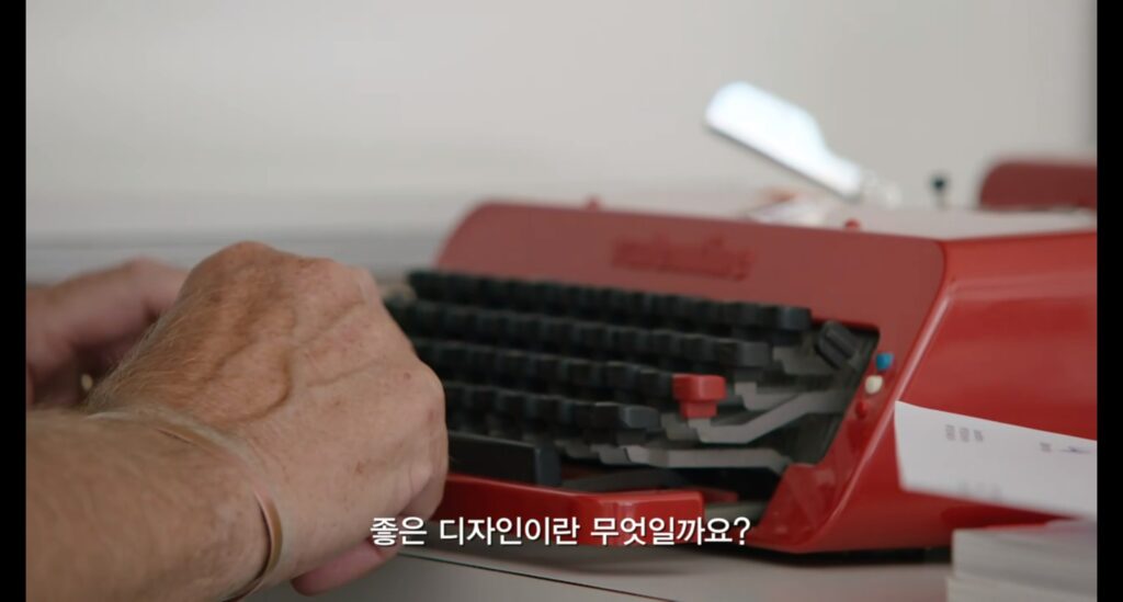 붉은색 타자기 위에 놓인 손과 하단에 '좋은 디자인이란 무엇일까요?'라는 자막이 있는 다큐멘터리 장면