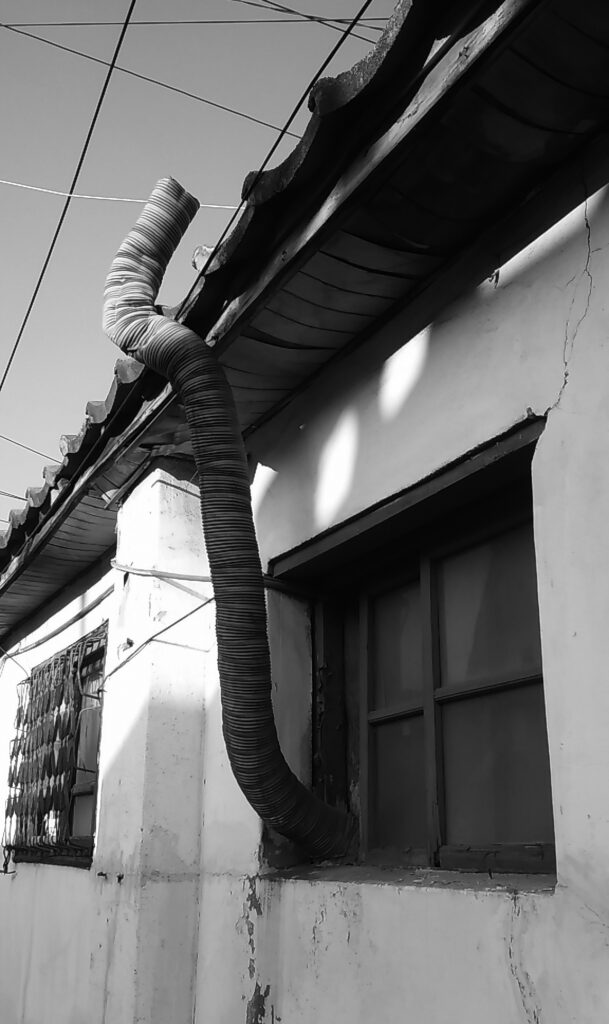 낡은 주택 외벽을 타고 지붕으로 올라가는 구불구불한 은색 연통(닥트)과 방범창이 있는 창문 흑백 사진
