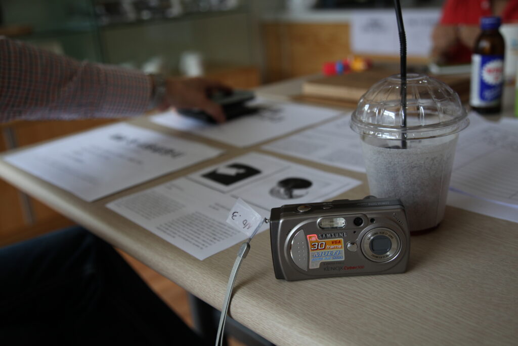 테이블 위에 놓인 삼성 디지털 카메라와 그 옆에 놓인 사물 인터뷰 기록지, 투명한 테이크아웃 컵