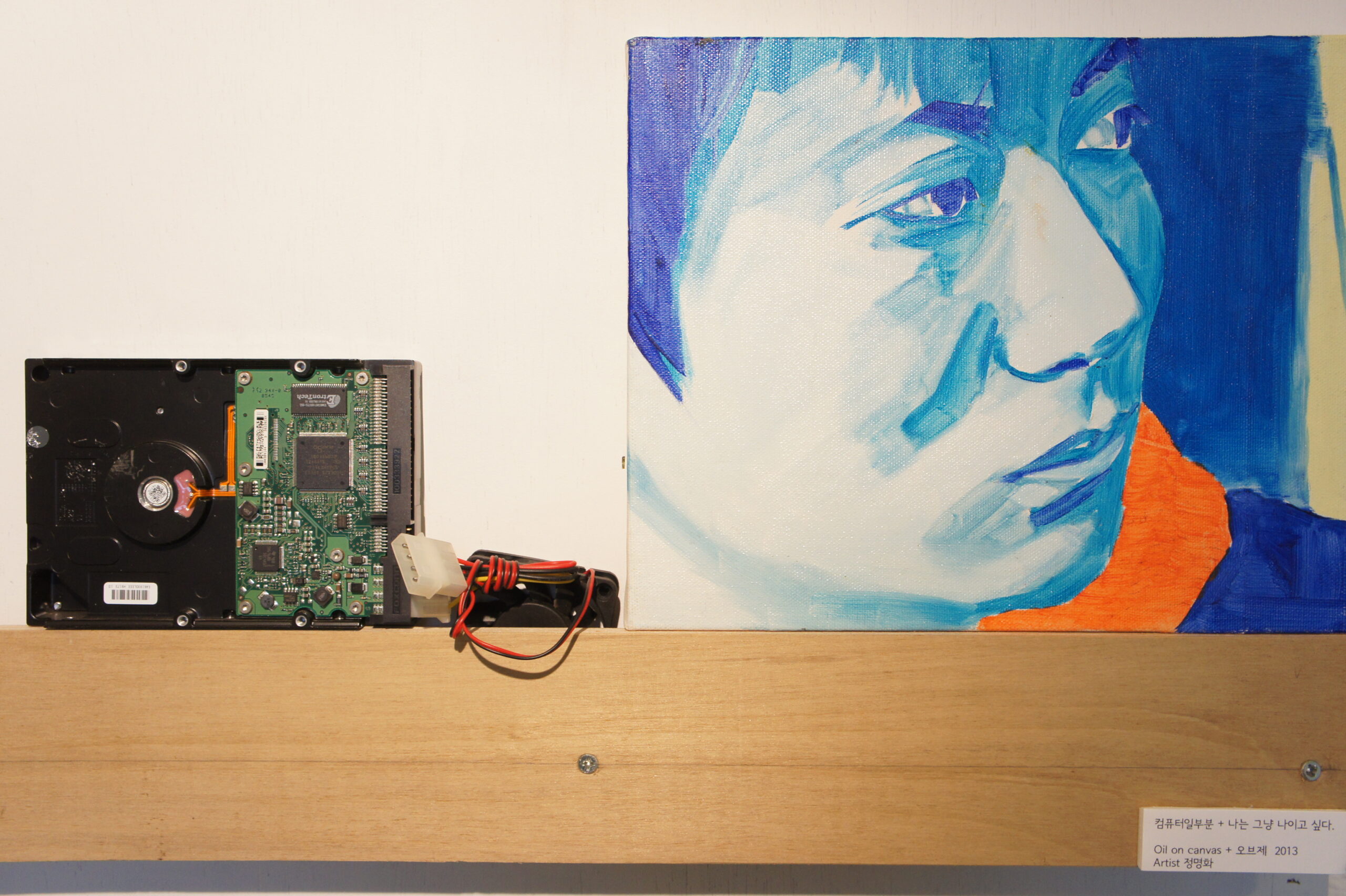 (정명화 작가 연작) 나무 선반 위에 놓인 컴퓨터 하드디스크 부품과 그 옆에 배치된 푸른색 톤의 인물 초상화 유화 작품.